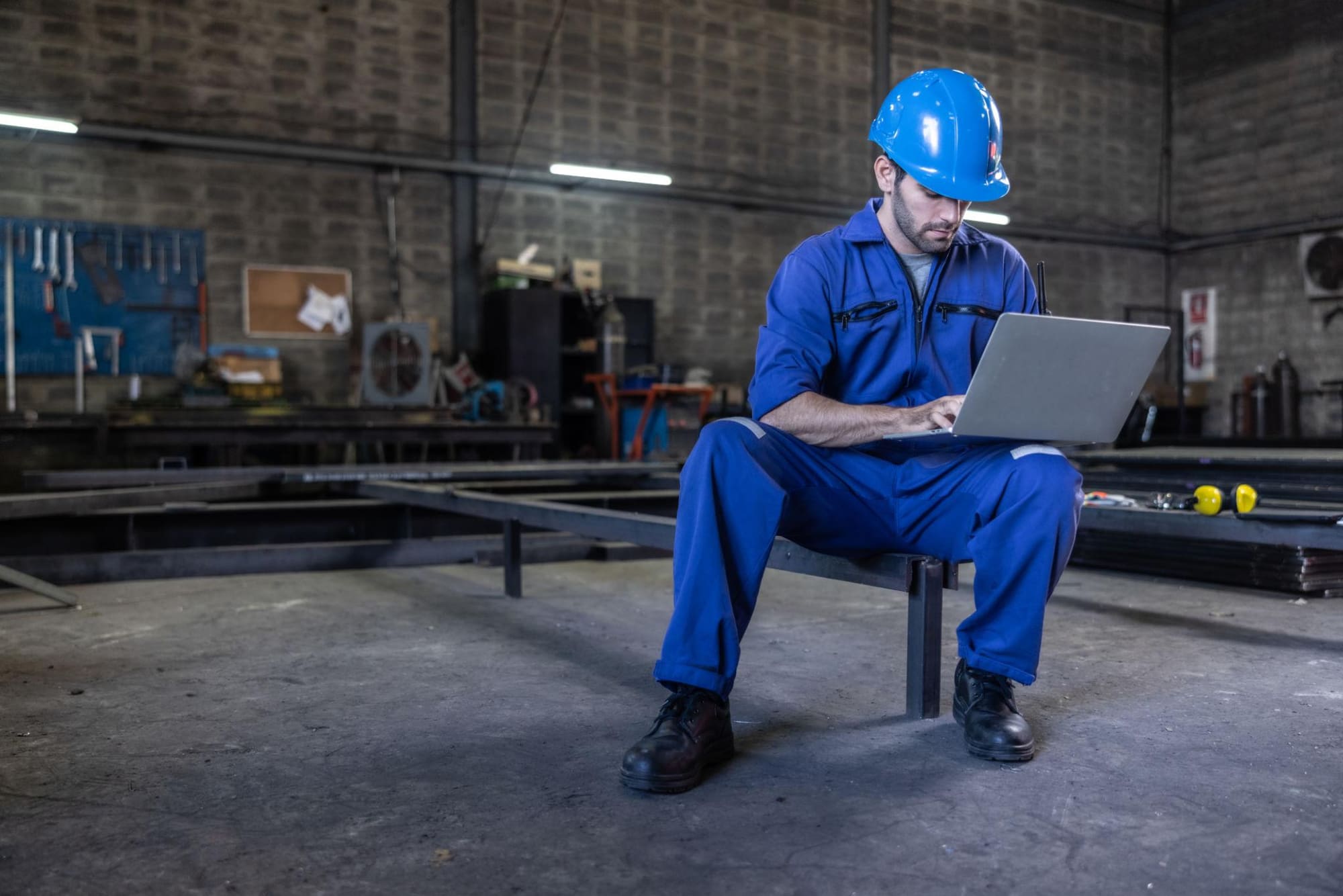 Machinist sitting on a workbench using a laptop in a fabrication shop
